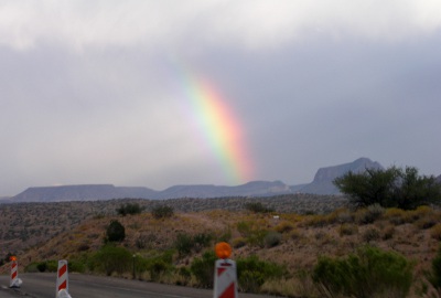 [Another rainbow arcing from upper left to lower right. Traffic barriers on the road seen in foreground.]