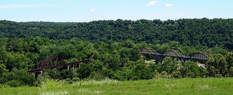 [A railroad bridge angles from the lower left towards the middle of the image while a road bridge angles from the lower right to the middle of the image. Both viewable sections of the bridges start and end in the greenery of trees so it's not possible to see from where they come or to where they go. The valley and nearby hillsides are full of leaf-bearing trees.]