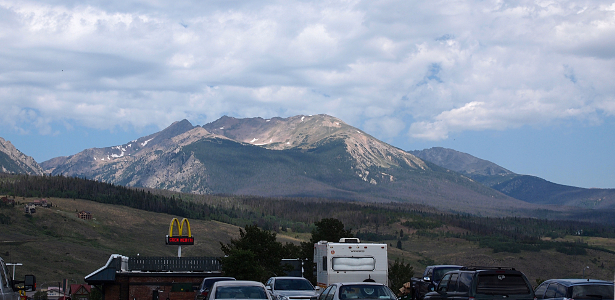 [Mountains with a scattering of snow rise in the distance above a parking lot full of vehicles in front of a McDonalds.]