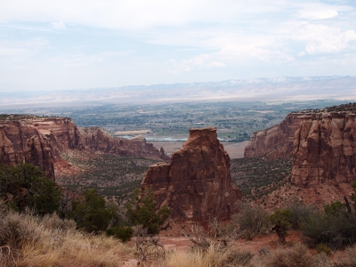 [A two-level free standing rock formation in the middle of a canyon. The top of this rock is about even with the canyon walls which can be seen on either side of it going into the background of the photo which opens to the town of Fruita.]