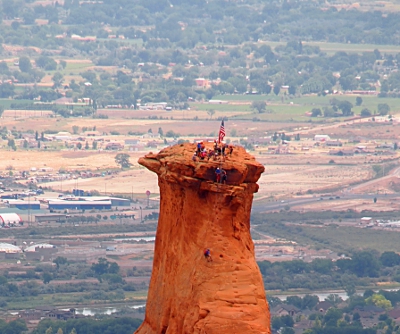 [Closeup view as seen from the skinny side. One person is either still climbing up or is rappelling down. Most people and the flag are at the top. Two people are one level down watching the person climbing.]