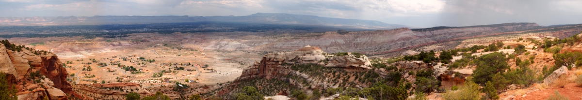 [Approximately 6 photos stitched together showing the vast expanse of canyons. On the left is a round section of land with specks of buildings and trees (only specks can be seen from this distance). The shades in the blue sky vary from light to dark depending upon the location of the rain.]