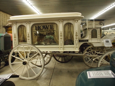 [All white hearse with gold inlay. Three large windows have heavy white drapes covering them. Hearse has four wooden wheels.]