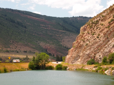 [The ramp and the interstate runs through a valley in the mountains. On the far side is a green mountain side and on the near side is rock carved to make room for the road.]