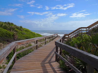 [A boardwalk built through the greenery-covered sand dune leads to waves seen in the distance. To the right a set of boardwalk steps lead up to a viewing platform. White puffy clouds are scattered through the blue sky.]