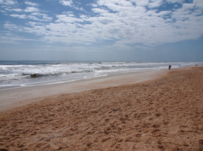 [An expanse of shell-laden sand with footprints all through it borders the smooth sand which meets othe ocean's waves. In the far distance are several lone people on the smooth sand. Many puffy white clouds cover the sky.]