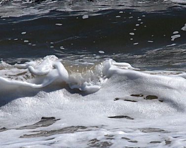 [The foreground is almost completely covered with teeny white bubbles. At the center of an image is a small section of water breaking over the bubbles and looks like a waterfall as the water is bubble-free. Behind this is still-looking dark blue water with a small scattering of white bubbles.]