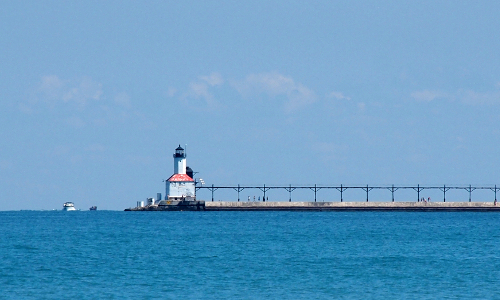 [Small lighthouse is left-center of photo in the water with a quay across the waterleading to it from the right. On the left of the lighthouse are several boats in the water.]