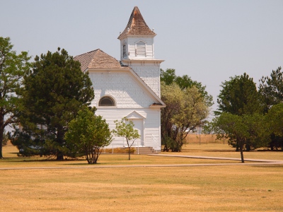 [White clapboard, two-story church with three story bell tower amidst some trees on the prairie.]