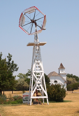 [White, wooden windmill tower atop which sits a spoked wheel with wooden blades attached side-by-side perpendicular to the outer frame of the spoked wheel.]