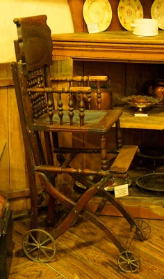 [Woodwn high chair with no tray. Front legs have small wheels while back legs have larger metal wheels.]