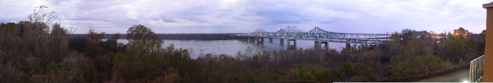 [Multiple images combined to create the scene. On the left are trees and vegetation. A silver bridge crosses the Mississippi River which appears calm under the cloud-covered sky. On the far right is the balcony railing and an edge of the brown-orange building.]