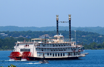 [Four level steam boat with two tall stacks and two red paddle wheels moving along the lake with several layers of treed hills in the background.]