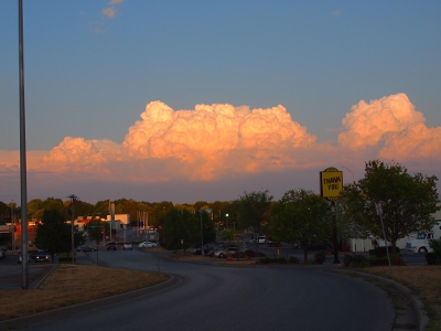 [In a clear blue sunset sky are thick, white clouds hanging above the street scene.]