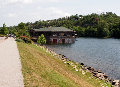 [Sidewalk at the top of the hillside slopes downward to the water. Ahead a restaurant with its supports in the lake sits aboe it.]