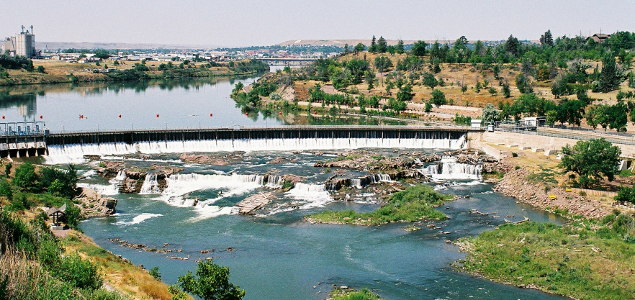 [The dam is a curved shape across the river. Downstream of the dam is a lot of rock sticking out of the water and creating several falls after the falls of the dam.]