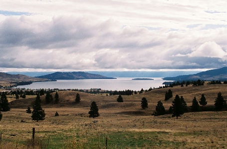 [Rolling hills with scattered short evergreens cover the ground near the lake. The sky is full of thick clouds which reflect on the water making it appear near-white rather than blue.]