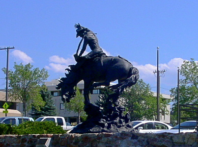 [The all-black statue sits on a stone wall. It is of a man on a bucking bronco with one hand holding the rope and the other in the air. There's a parking lot behind the wall and it's filled with vehicles.]