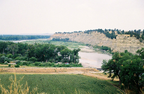 [There is a line of trees at the water's edge on this side of the river and behind is a farm field. On the other side of the river is a tall rock face--more a cliff--with trees at the top. The river is wide, but does not appear overly deep.]