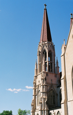 [It appears the top level of this spire has a bell. That level is open to the outside while the lower levels are close and appear to have stained windows. At the top of the spire's conical roof has a stone cross.]