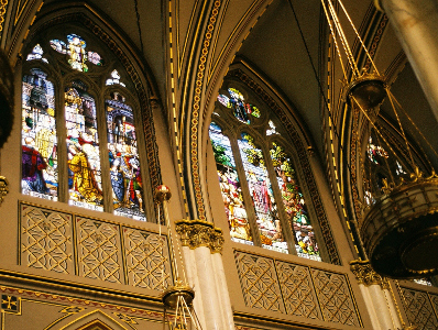 [Looking up at the stained glass panels on the second level. There are three panels under each arch depicting a scene. One of the gold chandeliers hanging from the ceiling is visible on the side.]