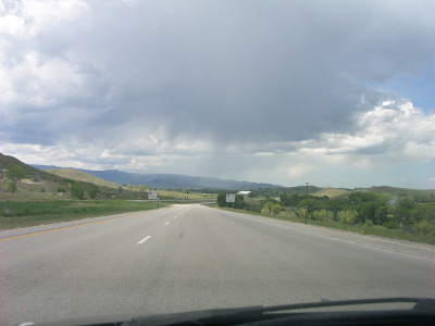 [Image taken through the windshield of a moving vehicle. There are no cars on the road ahead, but there is a dark cloud overhead that gets darker in the distance. No rain on the road in front of the car, but in the distance there are streaks of cloud going towards the ground.]