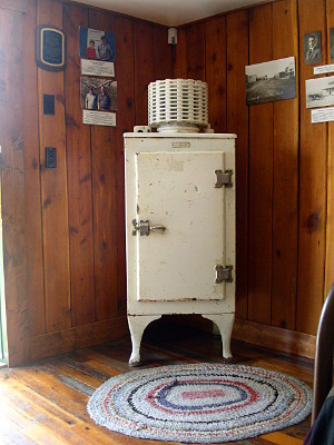 [An early refrigerator with a compressor on the top and the body rests on legs about a foot tall.]