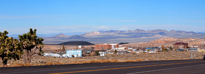 [Scenic roadside, panoramic view from the south end of the city. There are Joshua trees on the left at the roadside, colorful building/roofs sit on the dirt amid the brown sagebrush. In the background are mountains that appear to be colorful shades of white and purple due to shadows of clouds way overhead (and not seen in the blue sky of the photo).]