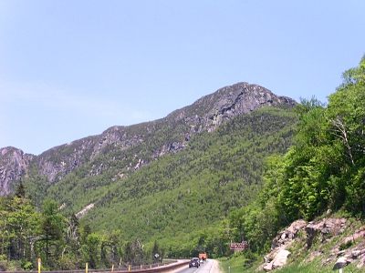 [Photo taken while driving of the whitish rocky mountain range with trees at lower levels towering over the two-lane roadway. with several vehicle a ways ahead of me.]