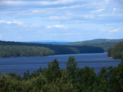 [Green trees in foreground, dark blue water in middle, layers of trees and several levels of mountains in background with haze showing the distance. Blue and white clouds in the sky.]