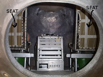 [An image taken through the oval opening showing a chair seat on either side with a silver box in between. The image was modified to add the word seat and an arrow to it for each seat.]