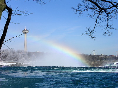 [This landscape view includes the tower on the left as the rainbow extends from the top of the falls to the buildings on the left. The rainbow colors are more distinct just above the water and fade as the bow gets closer to the buildings.]