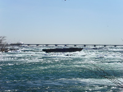 [A barge sits not moving in the choppy ice-laden water which is headed toward the falls. There are lots of whitecaps in the water, but the scow sits there like a rock.]