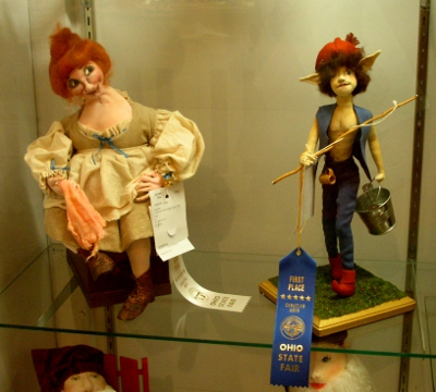 [Doll on the left with white ribbon hanging from it has red hair in a bun and wears a peasant dress. Doll on right with blue ribbon hanging from it is a caricature of the god Pan.]