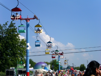 [Open lift cars (like at a ski resort) which pass over the fair grounds. People sit with feet dangling while others stand on the ground near food stands.]