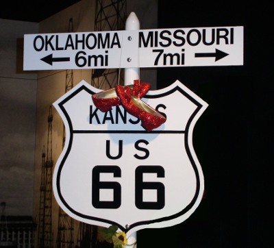 [Route 66 sign in Kansas with mileage to Missouri and to Oklahoma. A pair of sparkly red ladies shoes are tied together and hung over the Kansas part of the sign.]