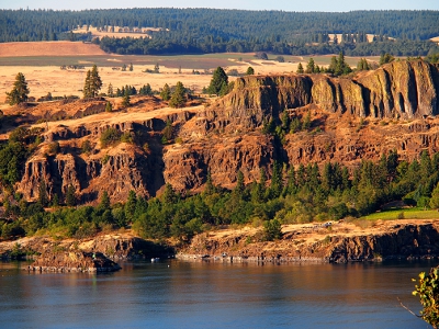 [Dark blue river in foreground and then a row of green evergreen and leaf-bearing trees and then several levels of gouged rock forming the hillside/cliffside. Flat fields and a carpet of evergreens can be seen in the distance.]