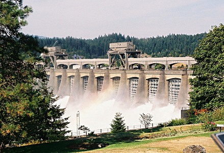 [The dam is a series of arched sections with whitewater coming through the gates. The spray of water creates a partial rainbow in it.]