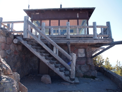 [Two level fire lookout station with first level made of large stones, stairwell to upper level and railing of upper level made of logs, and the top section nearly all glass.]