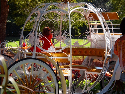 [White, metal, latticed, pumpkin shaped carriage with large white wheels and a golden seating area around the inside. One young girl stands on the seat looking back toward an elder.]