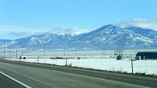 [The mountains have some snow at different levels on the mountains so there is white and grey going up the mountain side. The road is clear of snow, but there is a blanket of snow across the fields.]