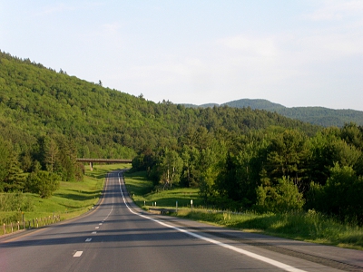 [A photo taken through the windshield of my car showing the two lanes of this half of the highway which curves off to the right in the distance. Mountains and plenty of trees are seen in the distance and along the road. Green grass is visible at the road edges.]