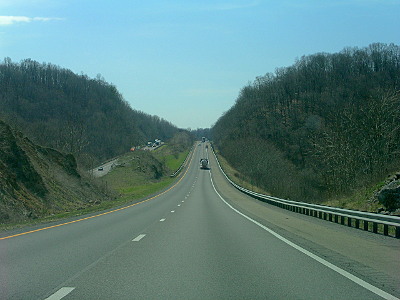 [View through the windshield of a vehicle looking at two lanes of highway with vehicles in the distance ahead. Along the right side is a hillside full of leafless trees. The highway has a hillside to the left on the divider between the northbound and southbound lanes which blocks at least half of the view of the lanes of opposing traffic. The sky is nearly all blue with a small section of white cloud in the upper left.]