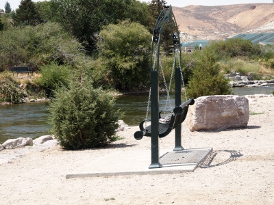 [The top of the back and the front of the seat are each linked by chains to the top of the overhead frame creating a swinging suspended bench which sits near the edge of the Green River. The frame and the bench are painted green.]